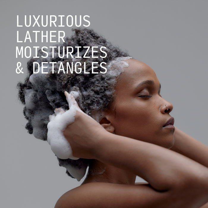 Model massaging Cécred Hydrating Shampoo into coily hair showing rich lather with Luxurious Lather Moisturizes & Detangles text overlay against a grey background