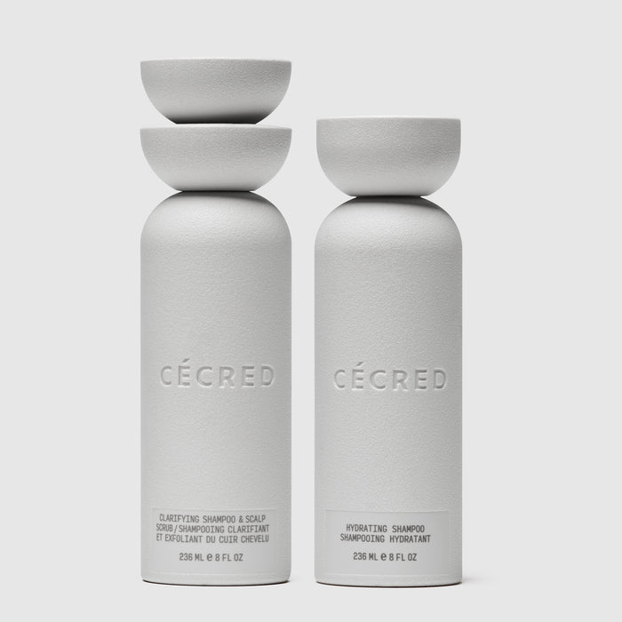 Two white cylindrical bottles labeled Cécred with matching bowls on top; left bottle is clarifying shampoo and scalp scrub, r