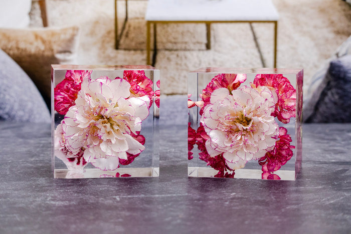 Two 3" clear resin keepsake blocks preserving pink and white carnations from a wedding bouquet, displayed on a gray table.