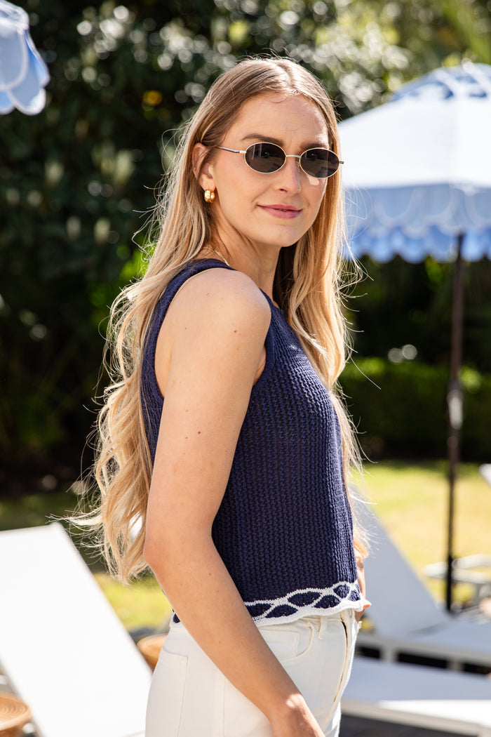 Woman wearing a navy blue sleeveless top and sunglasses outdoors with greenery and an umbrella in the background.