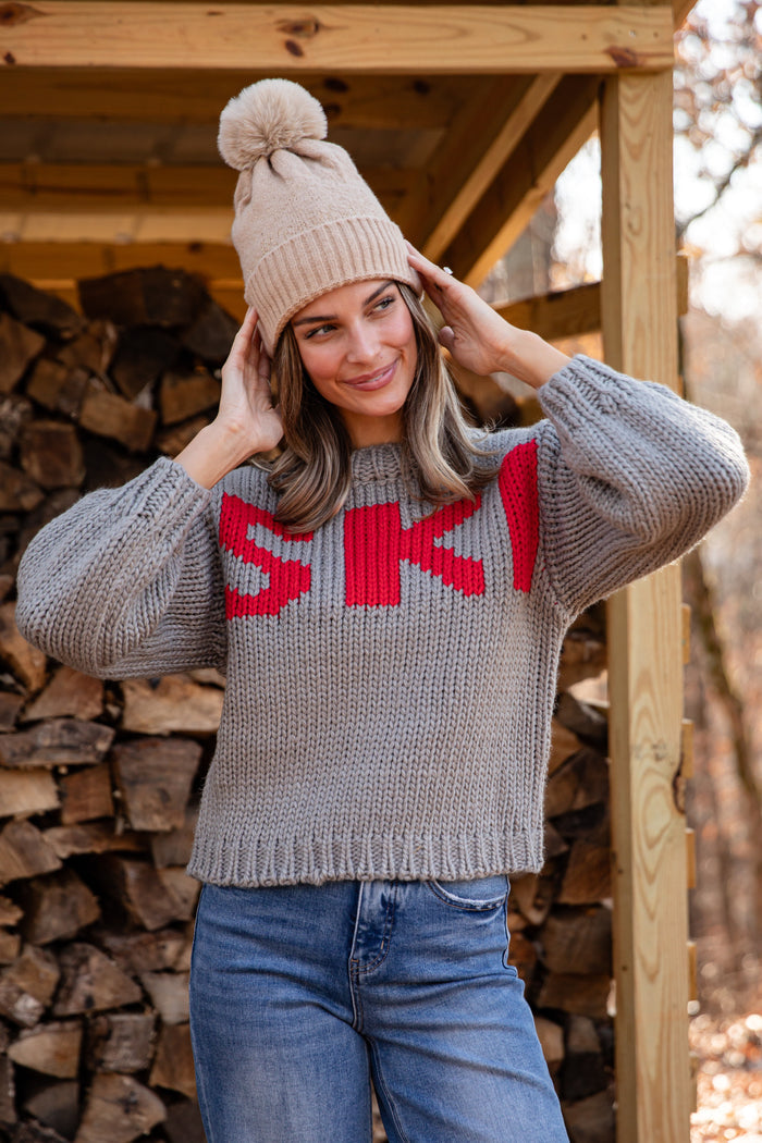 Woman wearing a gray sweater with 'SKI' and a beige knit hat, standing in front of stacked wood.