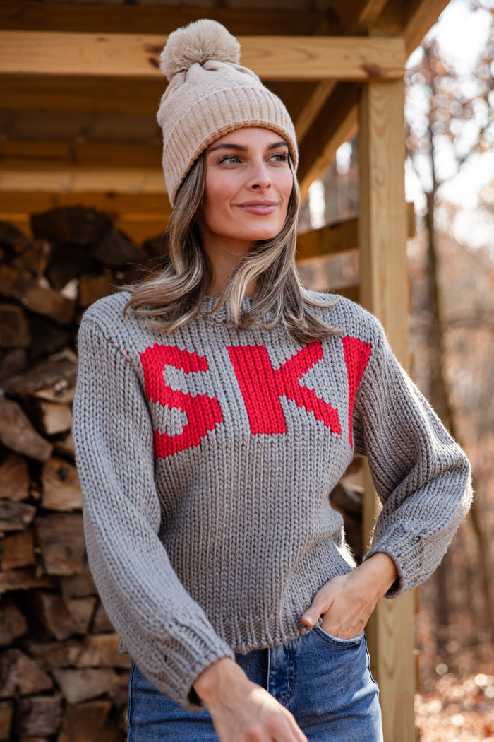 Woman wearing a gray sweater with 'SKI' in red letters and a beige knit hat, standing in front of stacked firewood.