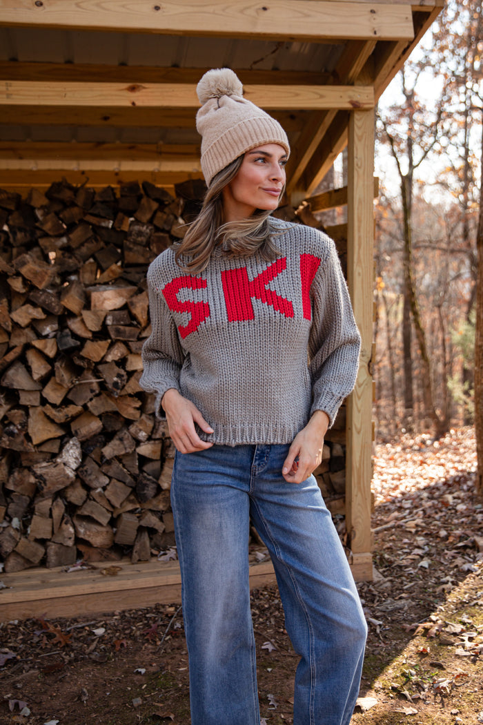 Woman wearing a gray sweater with 'SKI' in red, blue jeans, and a knit hat standing in front of stacked firewood.
