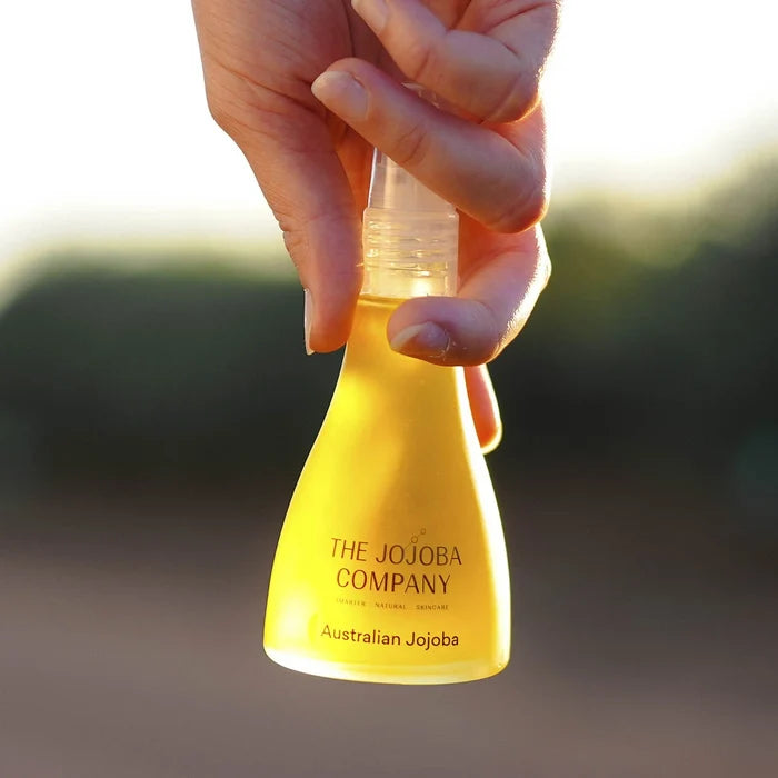 Hand holding a yellow bottle labeled 'The Jojoba Company Australian Jojoba' against a blurred natural background