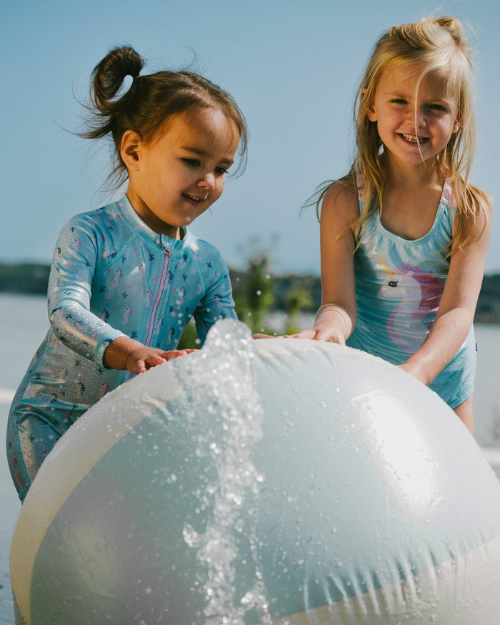 Side view of toddler girl in blue seahorse long sleeve rashguard swimsuit holding a beach ball