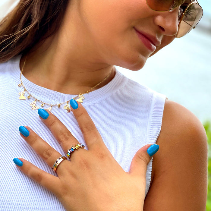 A woman in a white sleeveless top, wearing sunglasses, a butterfly necklace, and multiple rings. Her nails are painted vibrant neon blue with LONDONTOWN's Cabana Boy polish as she touches her chest with one hand, perhaps looking for Cabana Boy or waving to someone across the beach.
