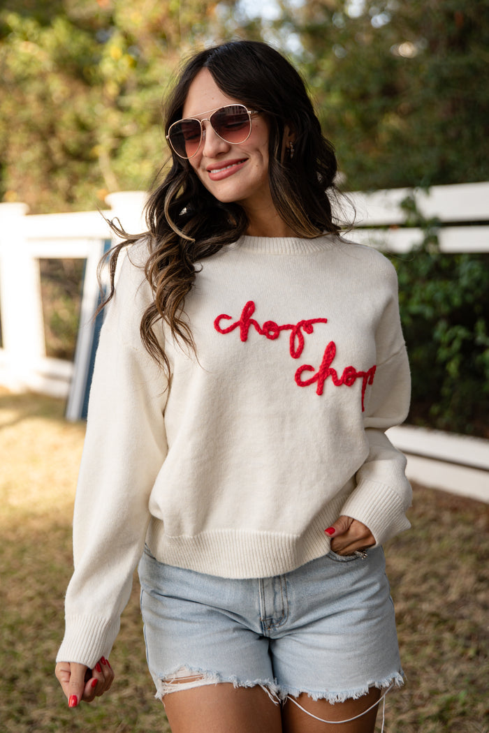 Woman wearing a white sweater with 'chop chop' text, sunglasses, and denim shorts outdoors.