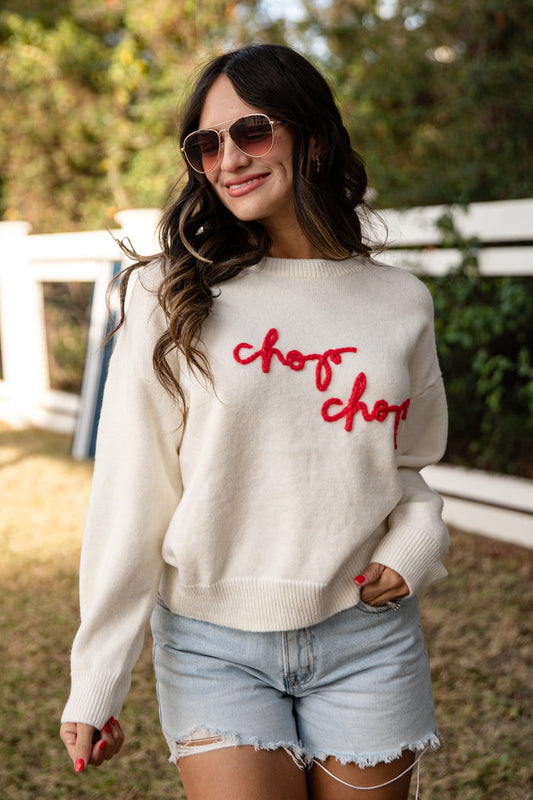 Woman wearing a white sweater with 'chop chop' text, sunglasses, and denim shorts outdoors.