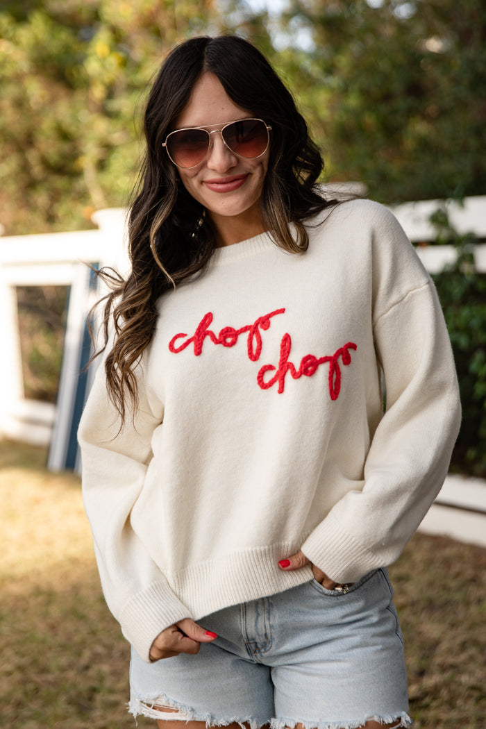 Woman wearing a cream sweater with 'chop chop' in red text, sunglasses, and denim shorts outdoors.
