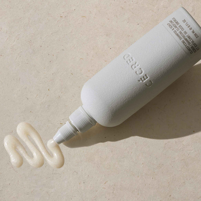 Cécred Clarifying Shampoo & Scalp Scrub bottle lying on its side with precision nozzle tip dispensing a swirl of creamy formula against a warm neutral textured surface