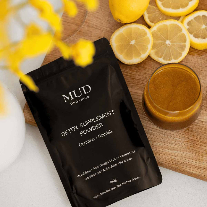 MUD Organics detox supplement powder package on a wooden surface with lemons and a glass of detox powder in water/juice.