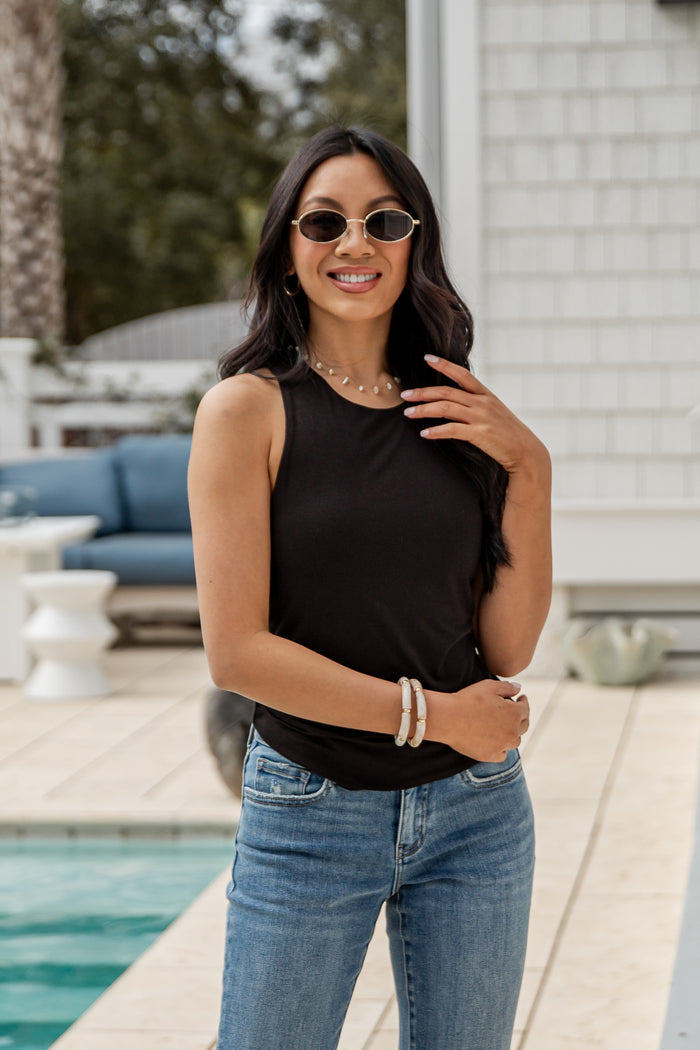 Woman wearing a black sleeveless top and blue jeans standing by a pool.