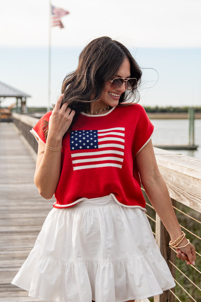 Woman wearing a red shirt with an American flag design and a white skirt on a wooden dock.