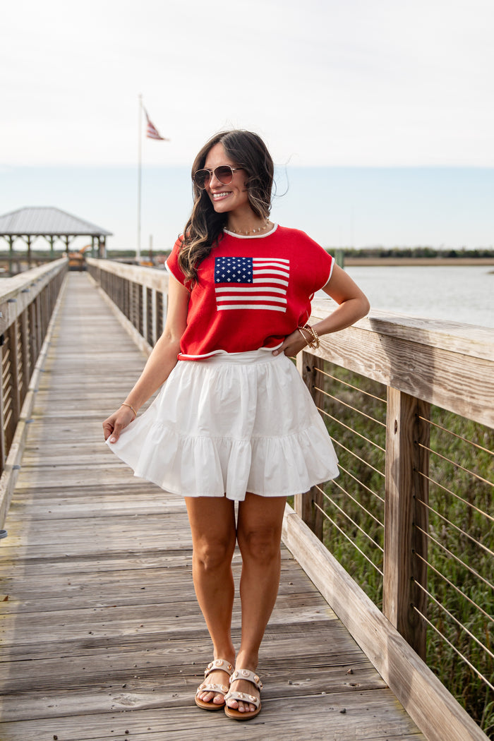 Woman wearing a red t-shirt with an American flag and a white skirt on a wooden dock.