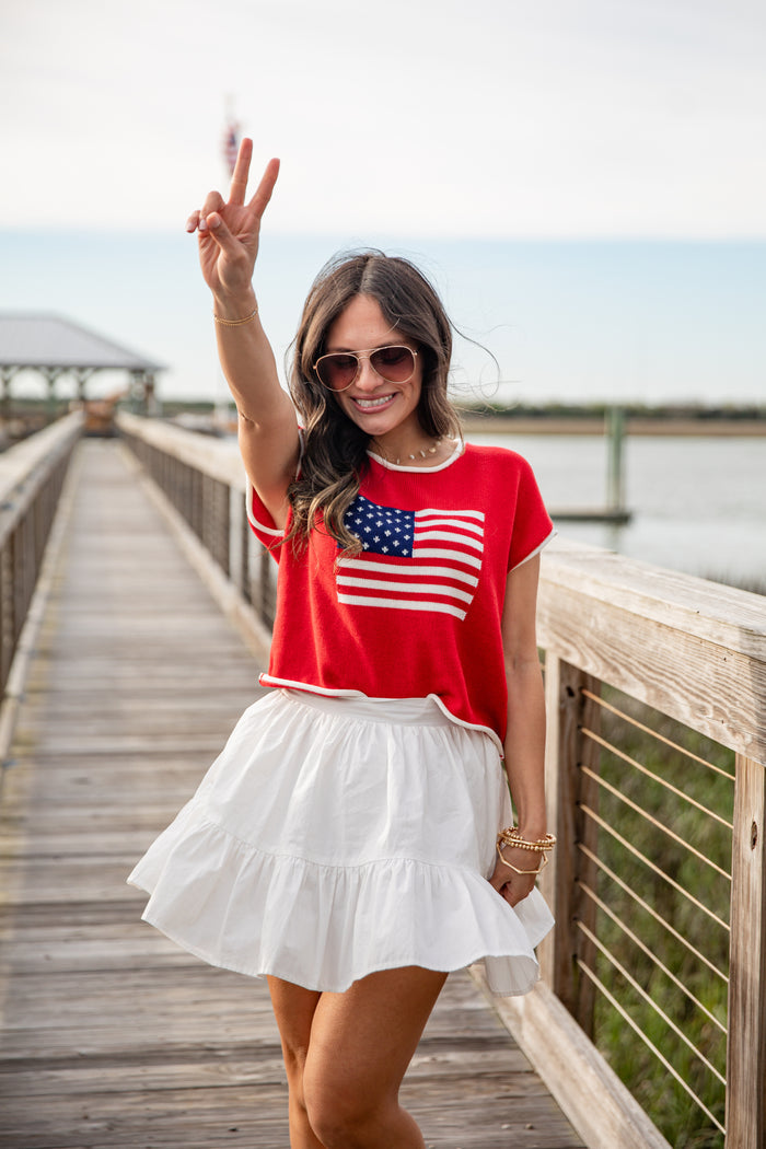 Woman wearing a red shirt with an American flag design and white skirt on a wooden boardwalk.