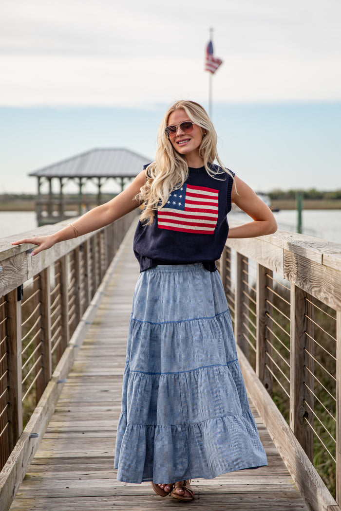 Woman wearing an American flag shirt and blue skirt on a wooden pier with a flag in the background.