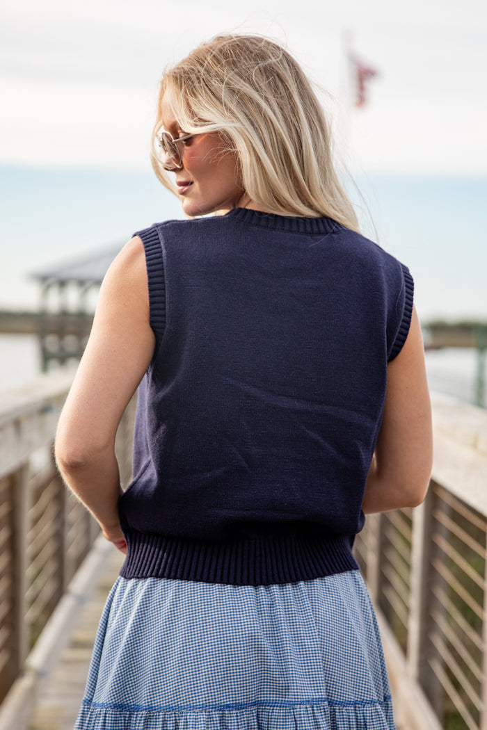 Woman wearing a navy sleeveless sweater and blue skirt on a wooden deck by a body of water.