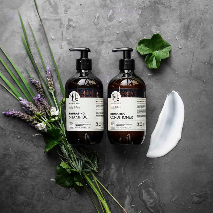 Two bottles of shampoo and conditioner with herbs and a leaf on a grey surface.