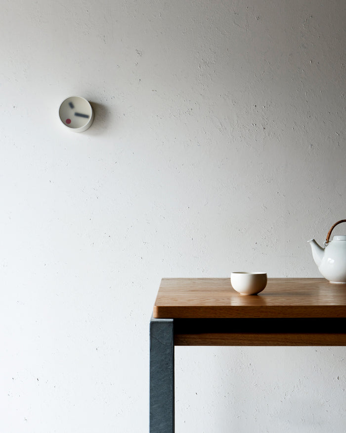 In situation mage of White Kehai Clock by Koizumi Studio on white wall next to wood table  with teacup and teapot.
