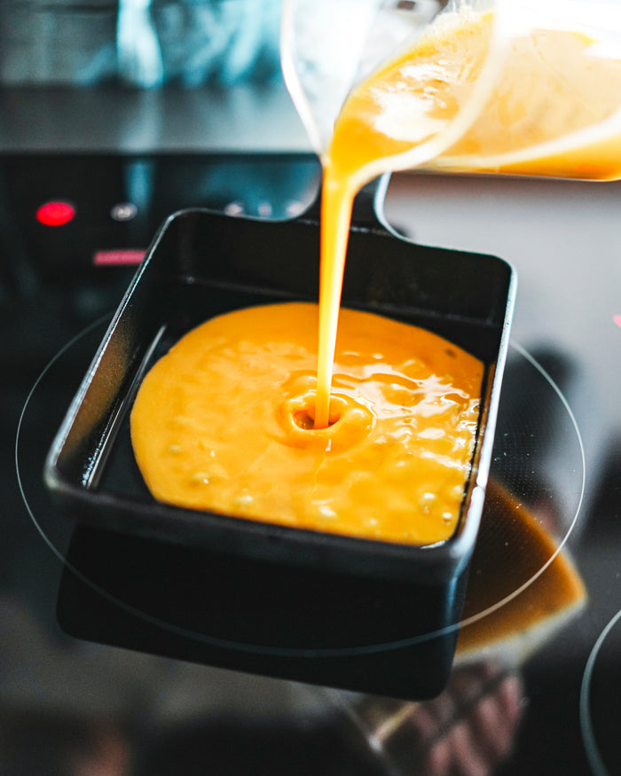 Pitcher pouring yellow egg mixture into a cast iron pan on a stovetop.
