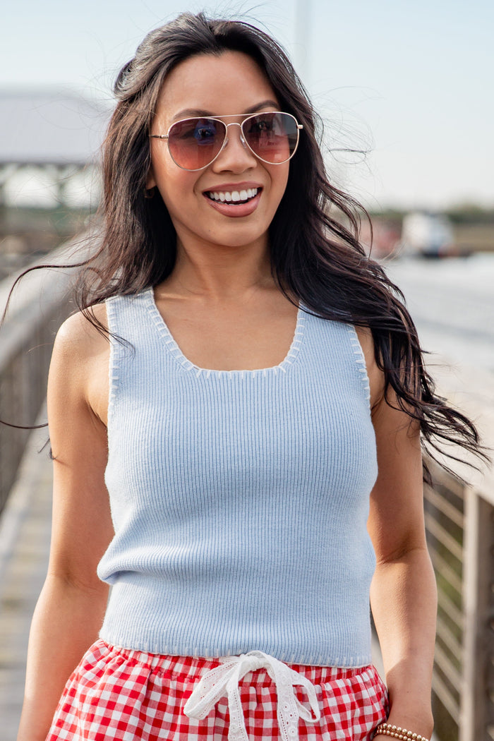 Woman wearing a light blue sleeveless top and red checkered shorts with sunglasses, standing outdoors.