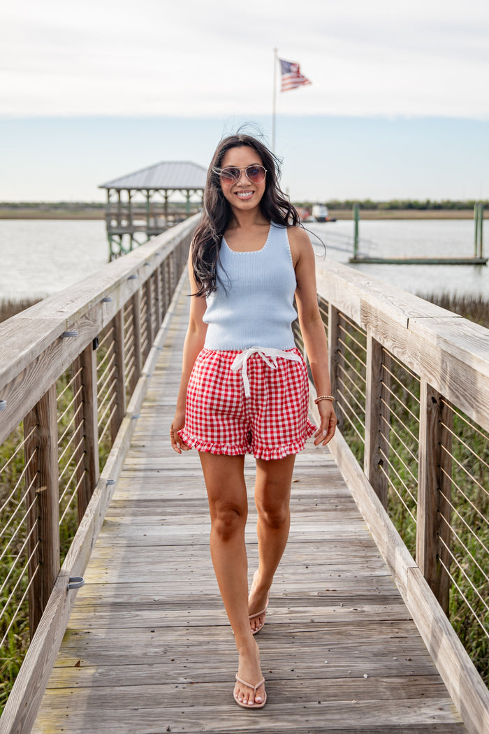 Woman walking on a wooden dock wearing a light blue sleeveless top and red checkered shorts.