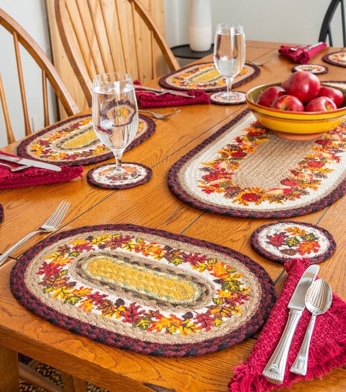 A dining table set with autumn-themed braided placemats with leaves design, matching coasters, and red fringed napkins, with a yellow bowl of red apples in the center.
