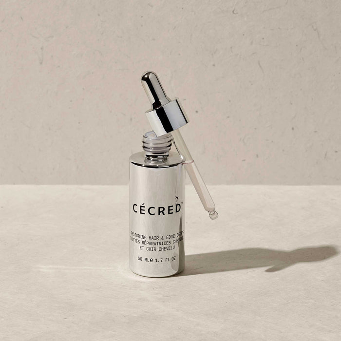 Cécred Restoring Hair & Edge Drops 50ml silver bottle with dropper removed and resting against the bottle on a warm beige surface, highlighting the precision applicator design