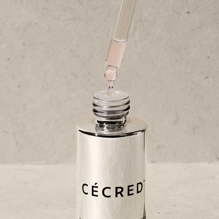 Extreme close-up of a Cécred Restoring Hair & Edge Drops dropper dispensing a single drop of clear serum into the open bottle, highlighting the formula's lightweight consistency