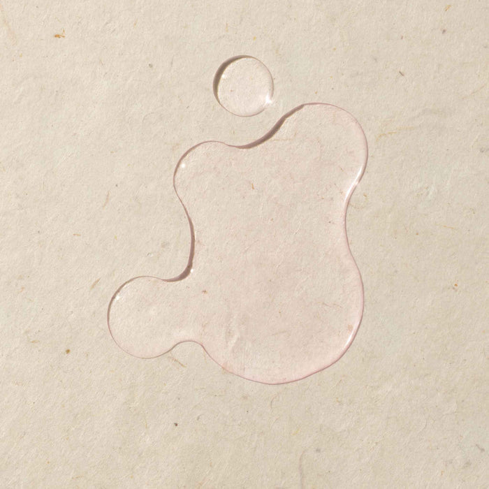 Close-up of a clear Cécred Restoring Hair & Edge Drops serum swatch on a warm beige surface, showcasing the product's lightweight, translucent pink fluid consistency