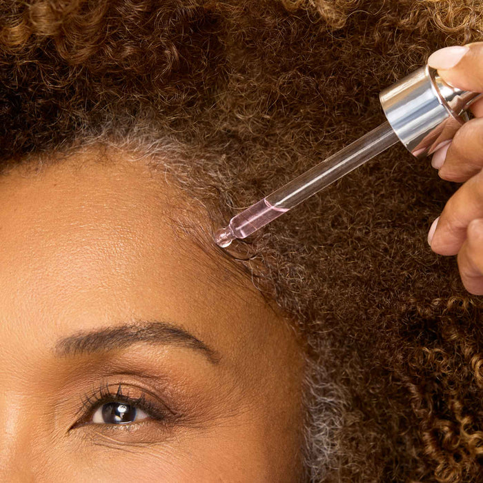 Close-up of a model applying Cécred Restoring Hair & Edge Drops dropper along her hairline, showcasing use on natural coily hair with gray highlights