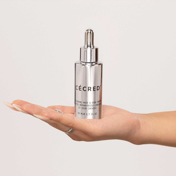 Hand holding a Cécred Restoring Hair & Edge Drops 50ml silver dropper bottle against a white background, highlighting the premium packaging and product scale