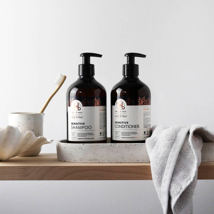 Two bottles of sensitive shampoo and conditioner on a wooden surface with a towel and bowl in the background.