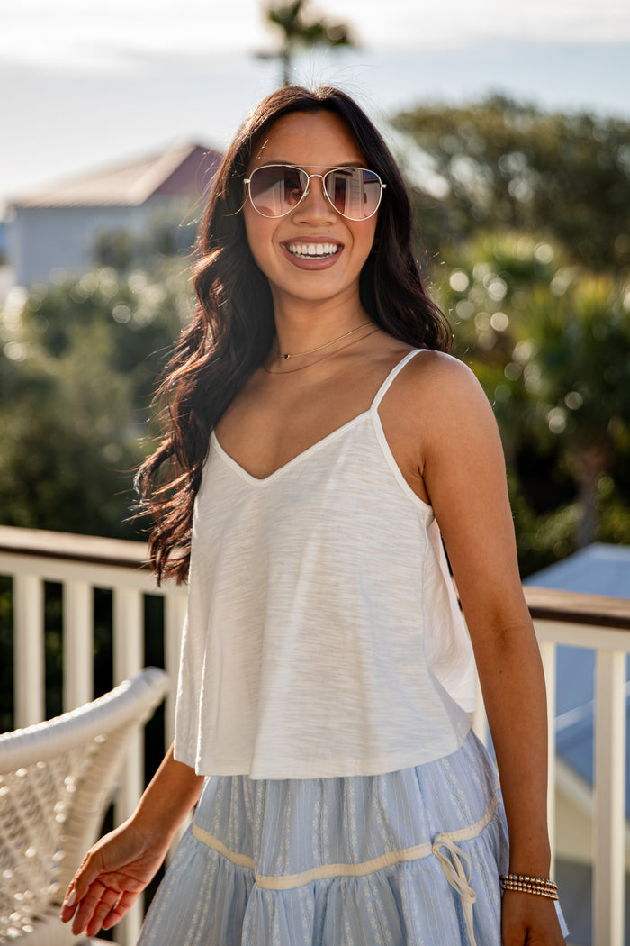 Woman wearing a white tank top with a blue skirt on a balcony with sunglasses and gold necklace
