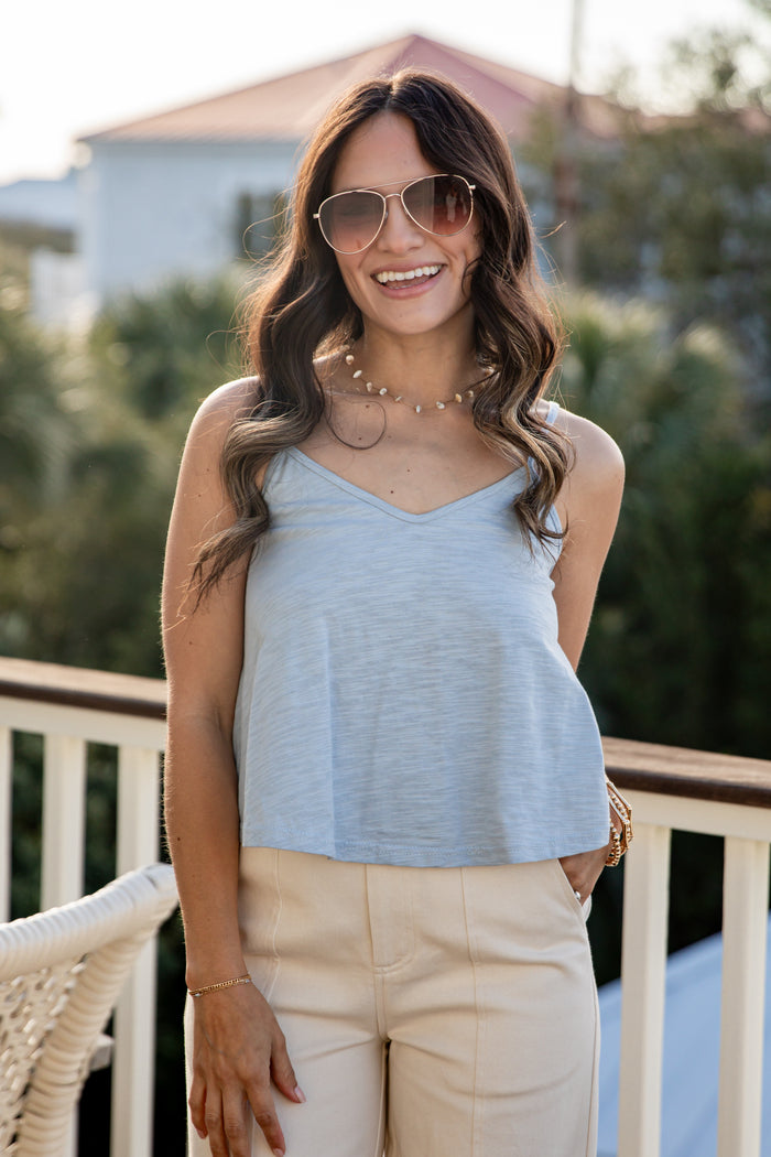 Woman wearing a light blue top and beige pants on a balcony with greenery in the background