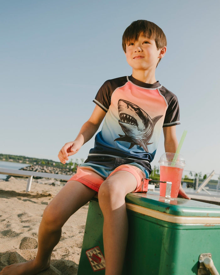 Boy sitting near the water wearing blue/orange gradient swim shorts and rashguard
