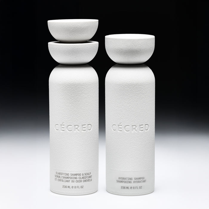 Two white cylindrical bottles with matching bowl-shaped caps labeled "Cécred," containing clarifying and hydrating shampoos.