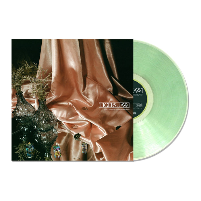 A limited edition green vinyl record by Tigers Jaw is partially pulled from its album cover, which displays metallic pink fabric, glass vases, and decorative objects arranged on a reflective surface.
