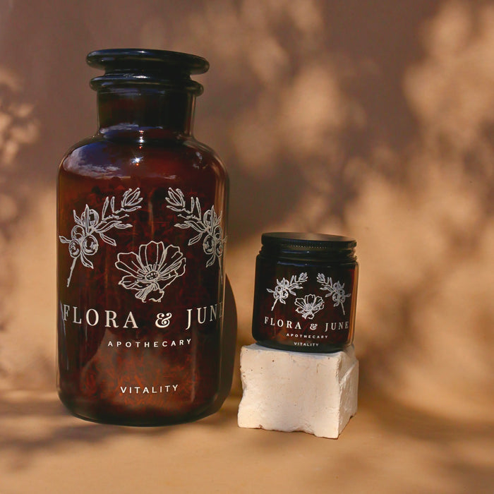 Two sizes of amber glass jars filled with Flora & June's Vitality loose leaf tea. Jars have a white label showing the brand name "Flora & June Apothecary" and the brand logo of a blooming flower and two sprigs of Juniper branch.
